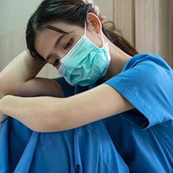 A nurse in scrubs wearing a face mask, who is sitting down and looking exhausted