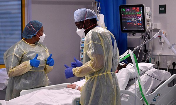 Two nurses at a patient's bedside in hospital, both wearing PPE
