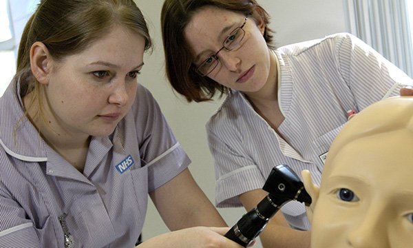 A nursing student holds an otoscope to the ear of a manikin as her trainer looks on