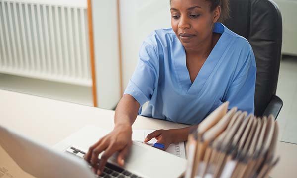 A nurse in blue scrubs sits at a desk using a laptop