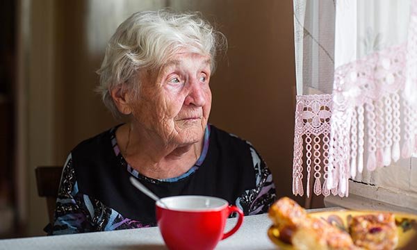 An older woman sits looking out of the window, seemingly disinterested in the plate of food and a cup in front of her