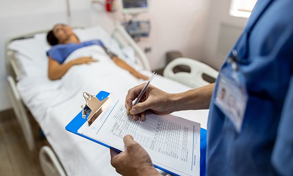 A nurse holding a clipboard makes notes while in the background a patient lies in a hospital bed