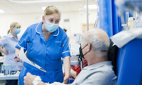 A nurse holds a pen and document as she talks to a patient sitting up in a hospital bed, both of them wearing protective masks